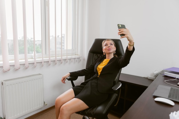 Relaxed business woman taking selfie on smartphone while sitting on boss's chair in office