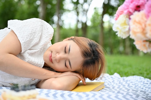 Relaxed and calm young Asian female laying down on a picnic blanket taking a nap in park