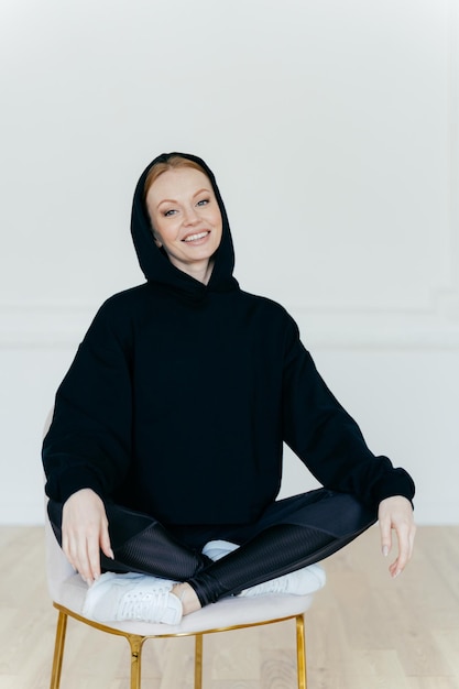 Relaxed cheerful woman in black sweatshirt sits in lotus pose on chair sits indoor has glad expression poses indoor satisfied after active training with coach People rest motivation concept