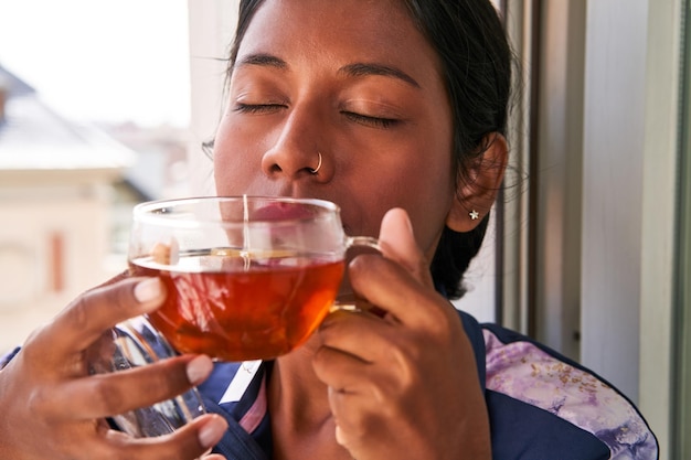 リラックスして居心地の良い若いインド人女性は窓辺で熱いお茶を楽しむ