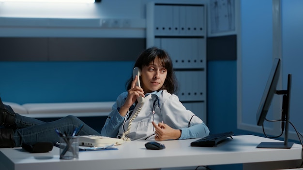 Relaxed doctor sitting with feet on desk table while talking at\
phone whith remote friend during night shift in hospital office.\
physician woman working at medical report analyzing expertise.