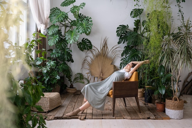 Relaxed dreamy woman in headphones listening music resting in surrounding of tropical plants at home