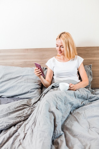 Relaxed european girl in white t shirt drinking coffee sitting on cozy bed. Indoor portrait of good looking happy blonde woman enjoying tea, reading text message on cell phone in morning.