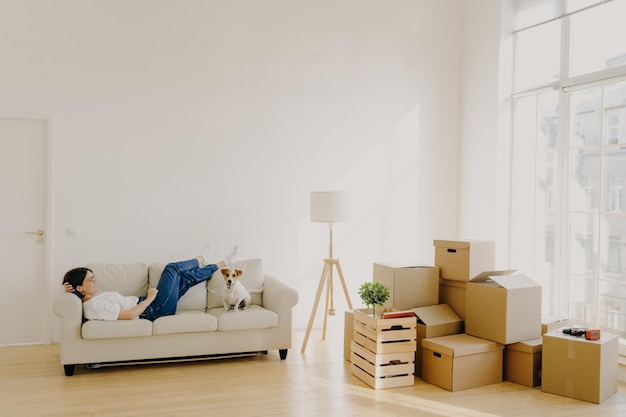Relaxed female lies on white comfortable sofa with dog feels tired after relocation and carrying boxes with belongings lamp stands near dressed in casual white t shirt jeans just arrives in house