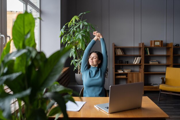 Photo relaxed female stretching hands taking break from work on laptop look in window in coworking space