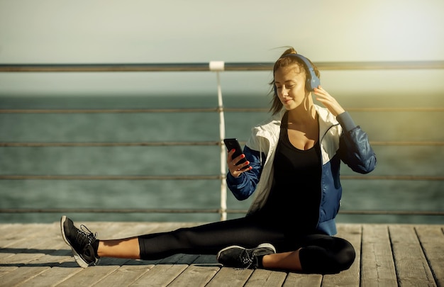 Relaxed fit woman in sportswear with headphones chooses music from a smartphone for training on the beach at bright sunny day