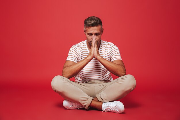 relaxed guy 30s in striped t-shirt sitting on floor with legs crossed and closed eyes isolated on red