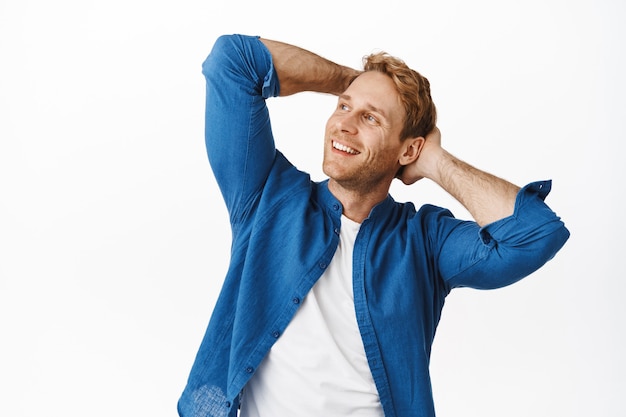 Relaxed handsome man holding hands behind head and enjoying leisure, weekend resting time, looking aside with chill smiling happy face, standing over white wall