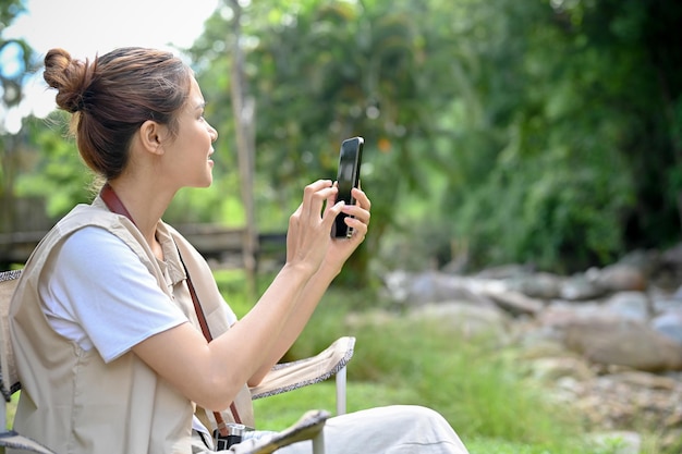 Campeggiatrice asiatica rilassata e felice seduta vicino al fiume e usando il suo smartphone