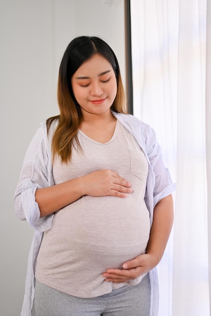 Relaxed and happy Asian pregnant woman in casual clothes feeling her baby touching her belly