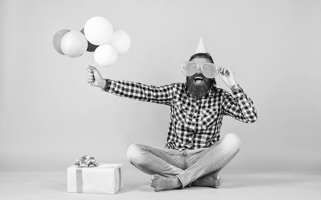 Расслабленный парень с днем рождения выглядит веселым бородатым мужчиной чувствует радость мужчина с подарочной коробкой празднует день рождения зрелый хипстер с яркими красочными воздушными шарами Сумасшедший забавный парень