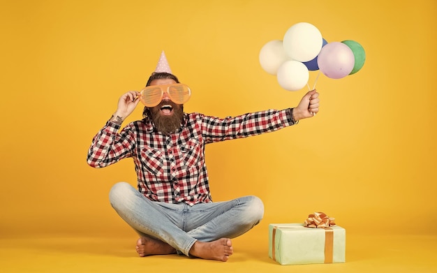 Relaxed happy birthday guy looks cheerful bearded man feel the joy man with gift box celebrating birthday mature hipster with bright colorful balloons Crazy funny guy