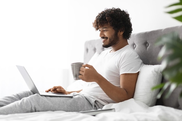 Relaxed indian man drinking coffee in bed using laptop