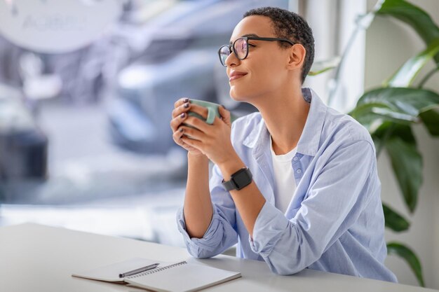 Photo relaxed lady in glasses savoring warm beverage with serene smile and notebook on table