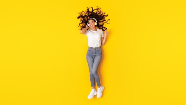 Relaxed lady singing listening music wearing headphones over yellow background