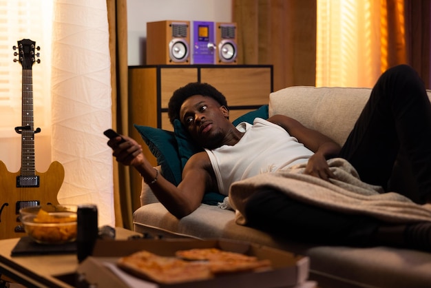 A relaxed man lies on the couch pizza and chips on the table next to him turns on the tv