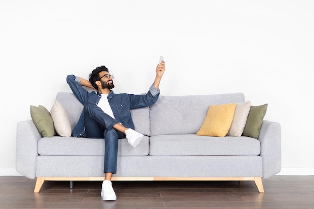 Relaxed middle eastern young man sitting on couch holding remote