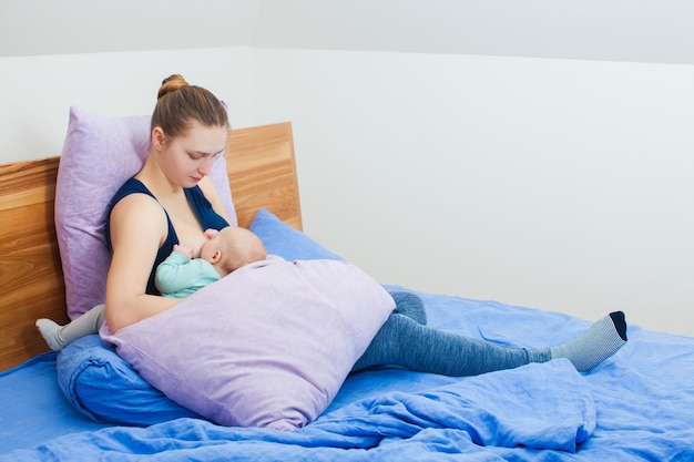 Relaxed mother breastfeeding infant baby sitting on bed in a bedroom surrounded by pillows that support her body and help to latch properly