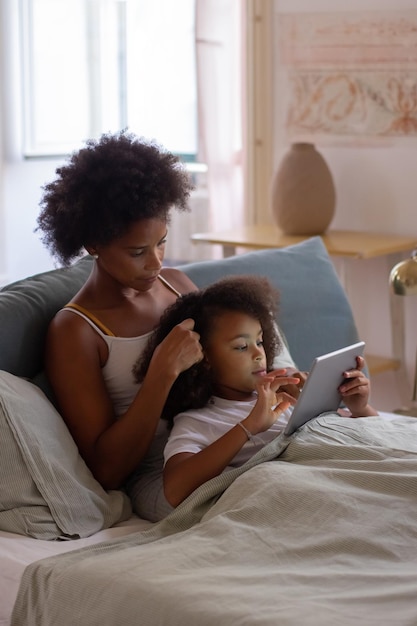 Расслабленная мать и дочь лежат в постели. Афроамериканская девочка с помощью планшета, мать гладит ее волосы. Семья, отцовство, технологическая концепция