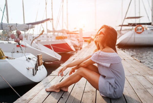 Relaxed and peaceful young woman sits on pier. She keeps legs and hands together. She wears sunglasses and striped dress. Young woman pose on camera.