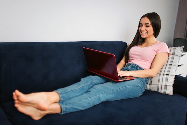 Relaxed and positive young woman sit on blue sofa in room. She smiles and type on laptop keyboard. Model work at home.