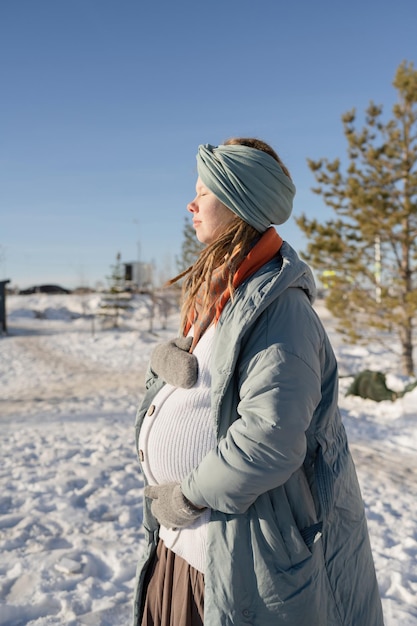 A relaxed pregnant young woman with her eyes closed, hugging her stomach in the park in winter