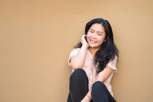 Relaxed Pretty Asian Woman sits on the rooftop floor with a light brown background and smiles to the camera