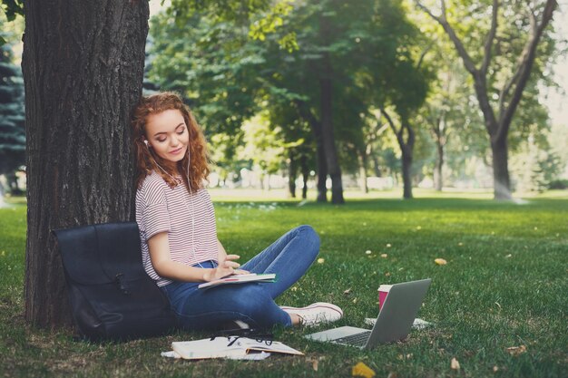 Relaxed redhead student girl listening to music online on smartphone in earphones sitting under a tree in the park. Education concept