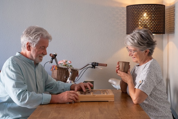 Relaxed senior couple spend time together at home playing a game of checkers on wooden table.