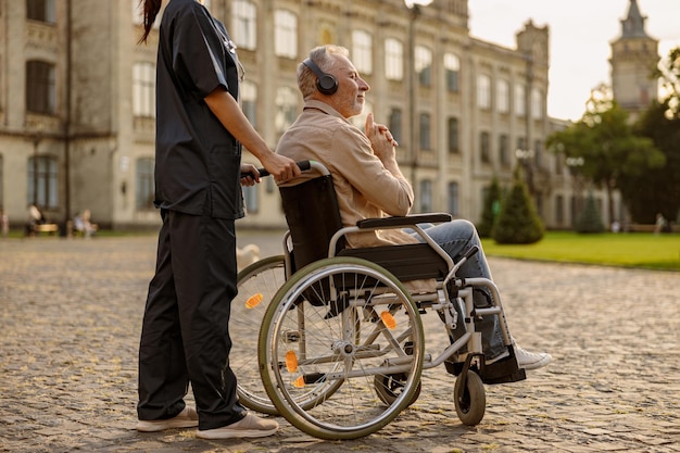 Uomo disabile anziano rilassato in sedia a rotelle che indossa le cuffie e ascolta la musica durante una passeggiata