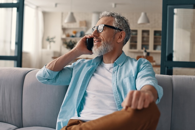 Uomo anziano rilassato in abbigliamento casual e occhiali che parla allo smartphone mentre è seduto sul divano di casa