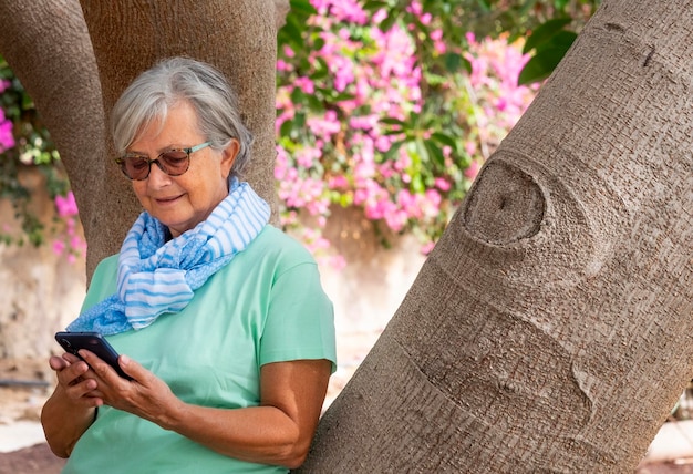 Rilassata donna anziana dai capelli bianchi che indossa occhiali da sole e sciarpa alla moda utilizzando smartphone e sorridente in piedi nel parco pubblico contro un tronco d'albero con bellissimi fiori rosa