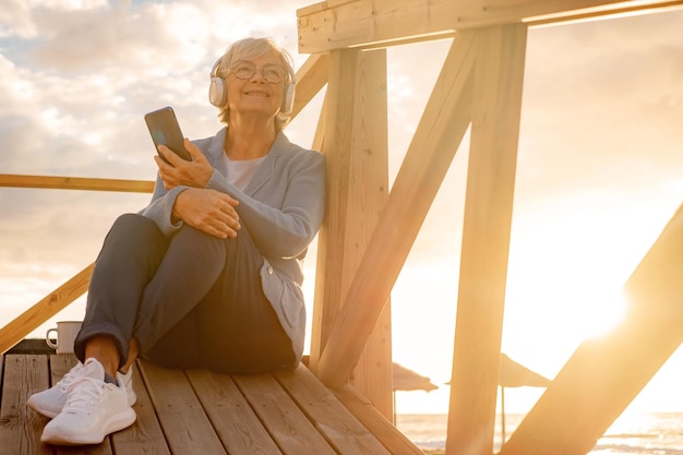 Relaxed senior woman with headphones sitting outdoors in sunset light listening music by phone