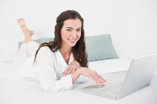 편안한 캐주얼 갈색 머리 침대에서 노트북을 사용하여 미소