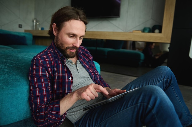 Расслабленный улыбающийся кавказец в повседневных джинсах и клетчатой рубашке читает новости на цифровом планшете, просматривает интернет, сидя дома на полу, откинувшись на диване.