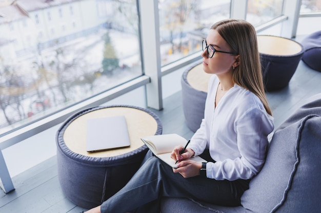 Relaxed smiling happy young woman in glasses and white shirt sitting in bean bag chair resting using laptop work making notes in notepad computer remote work concept