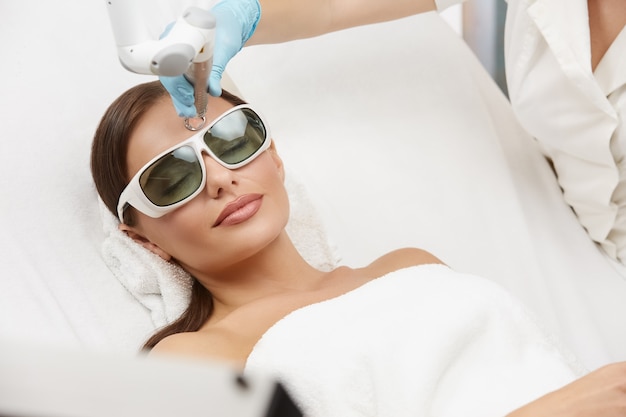 Relaxed woman receiving laser procedures in beauty salon on white bed, female getting facial treatment in luxury spa