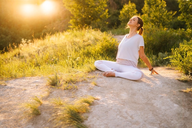 Relaxed woman sitting in lotus pose and doing bend in back outside in park evening on background of sunlight Female practicing yoga exercises in alone outdoors at green grass during sunset