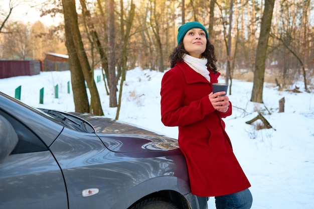 Relaxed woman traveler in warm winter clothes warming her hands holding a thermo mug with hot tea drink sitting on the car hood Sunbeams fall through pine trees on the snow covered forest path