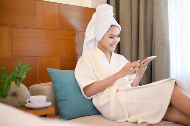 A relaxed woman wearing white bathrobe and towel having a coffee while using tablet in the morning