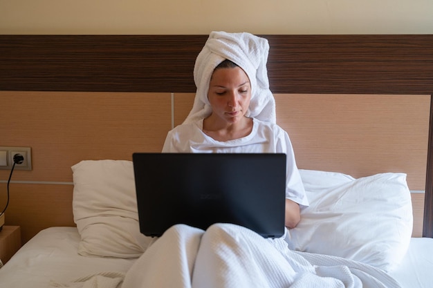 A relaxed woman in a white tshirt with a towel on her head is working on a laptop doing online shopping