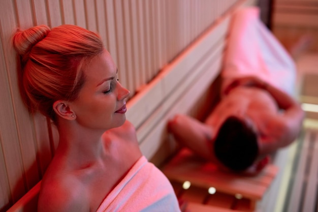 Relaxed woman wrapped in a towel relaxing in sauna at health spa Her husband in lying down in the background