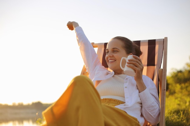 Relaxed young adult happy satisfied Caucasian female camper sitting on a camping chair and having a morning coffee near the river Camping and outdoor activity