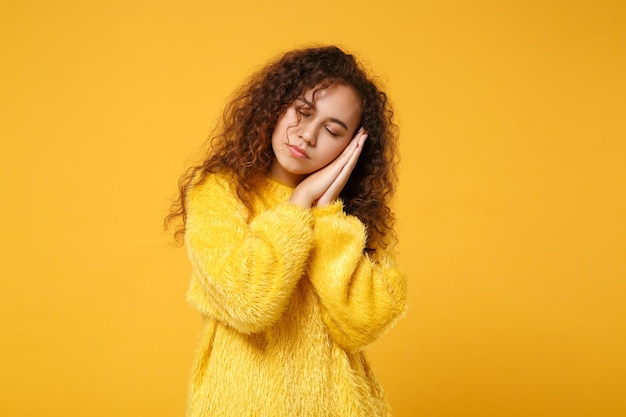 Relaxed young african american girl in fur sweater posing isolated on yellow orange wall background, studio portrait. People lifestyle concept. Mock up copy space. Sleep with folded hands under cheek.