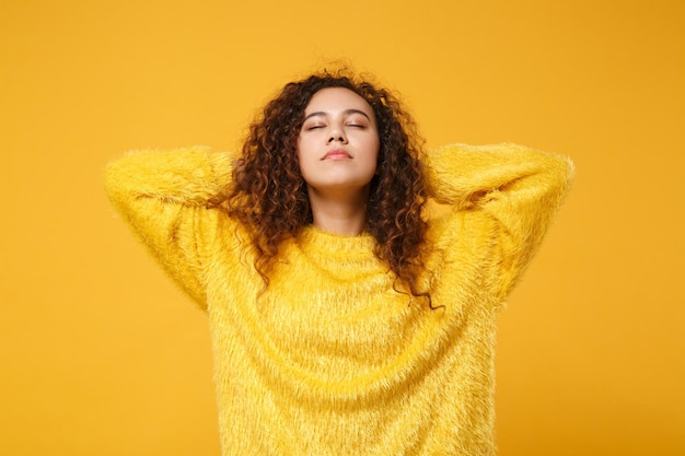 Relaxed young african american girl in fur sweater posing isolated on yellow orange wall background, studio portrait. people lifestyle concept. mock up copy space. sleeping with hands behind head