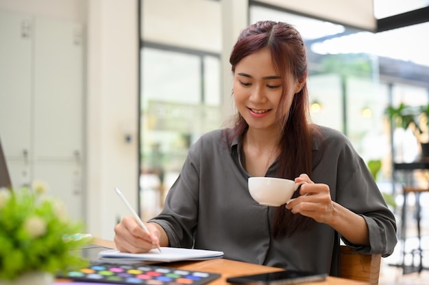 リラックスした若いアジアの女性のグラフィックデザイナーは、コーヒーブレイク中にコーヒーを飲みながら楽しんでいます