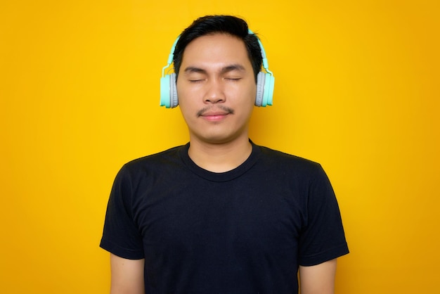 Relaxed young Asian man in casual tshirt listening music in headphones isolated on yellow background People emotions lifestyle concept