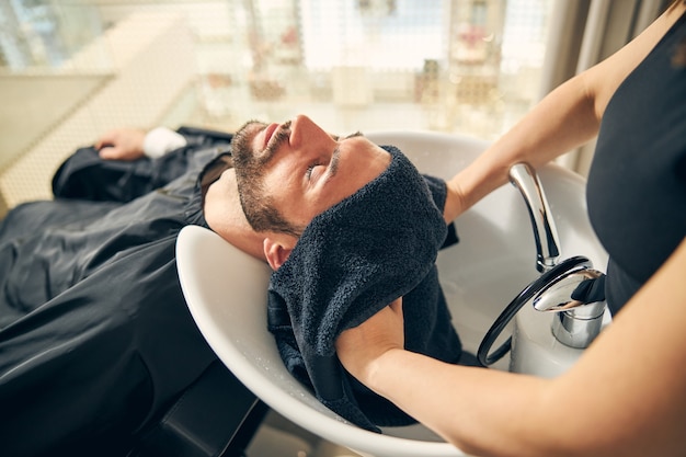 Photo relaxed young bearded man closing eyes while leaning on sink during procedure