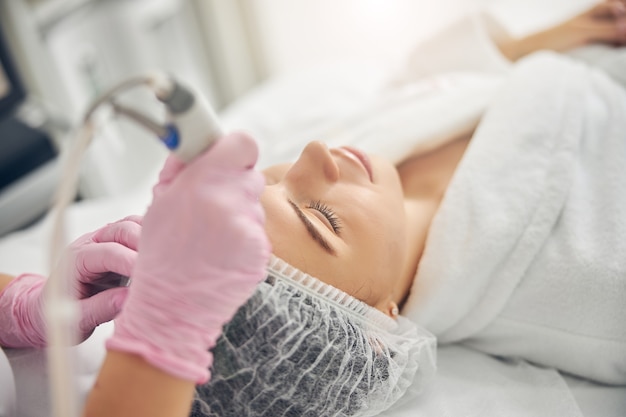 Relaxed young Caucasian woman in a bathrobe and a gauze cap during the beauty procedure