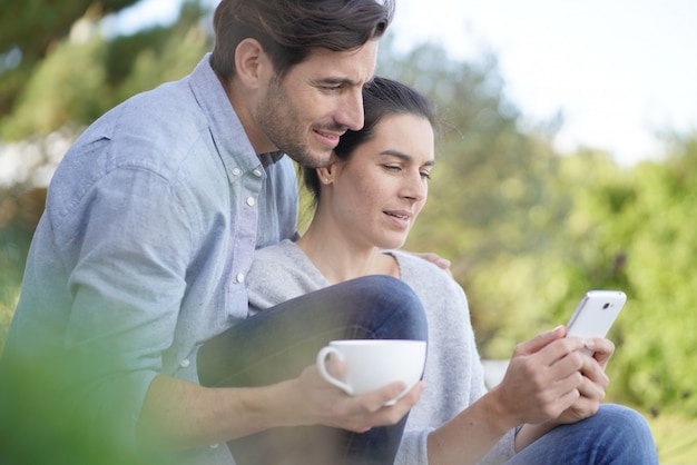  Relaxed young couple sitting together outside with mug and cellphone                                              